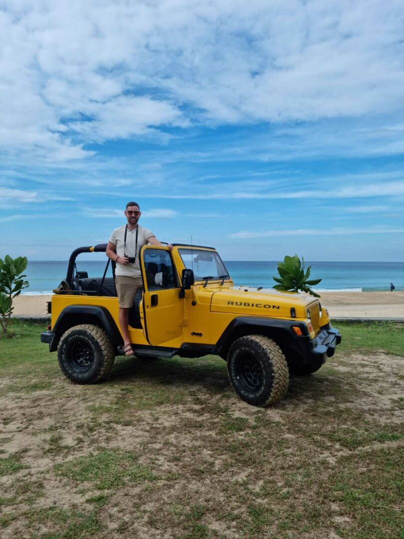 A man standing at the open door of a yellow Jeep Rubicon with a view of the ocean in the background.