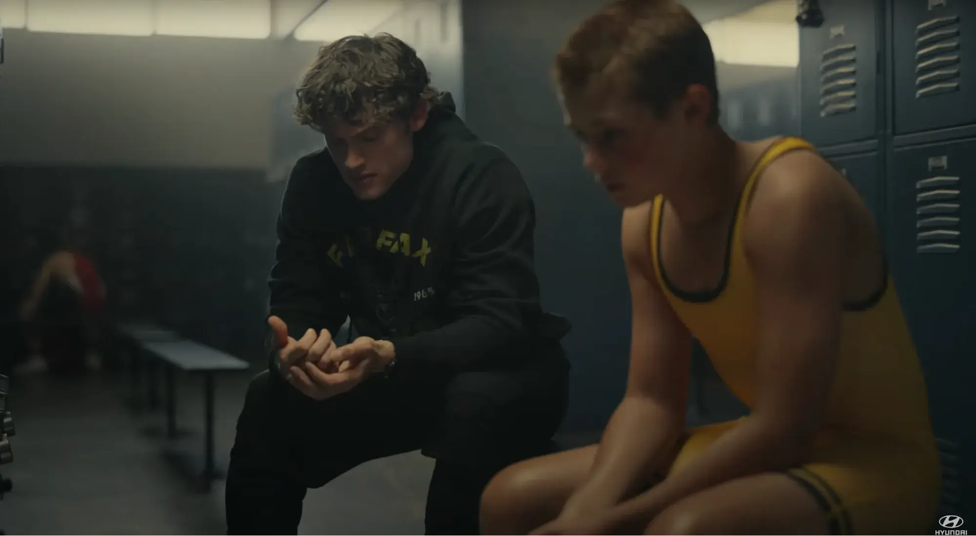 Screen cap from ad. A 20-something man sits next to a younger boy in wrestling gear in a locker room, having a serious conversation.